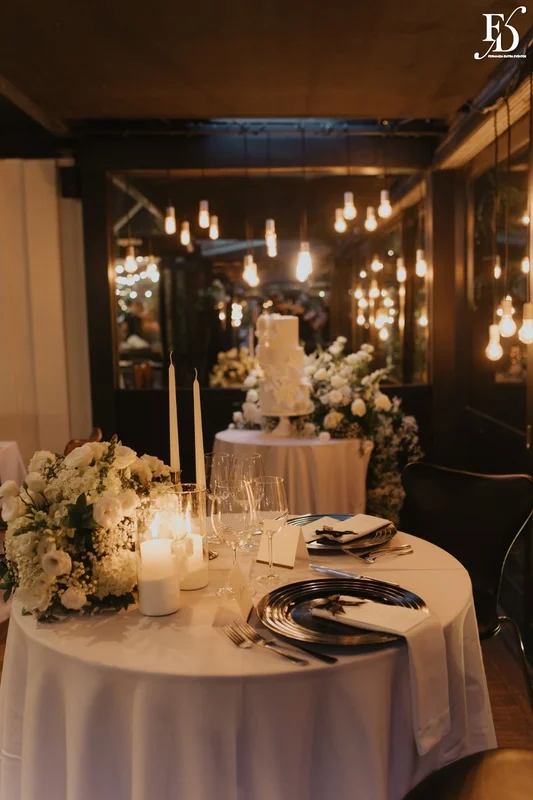 Mesa das noivas com arranjo orgânico branco, velas e bolo de casamento ao fundo no Le Bistrot Gourmet