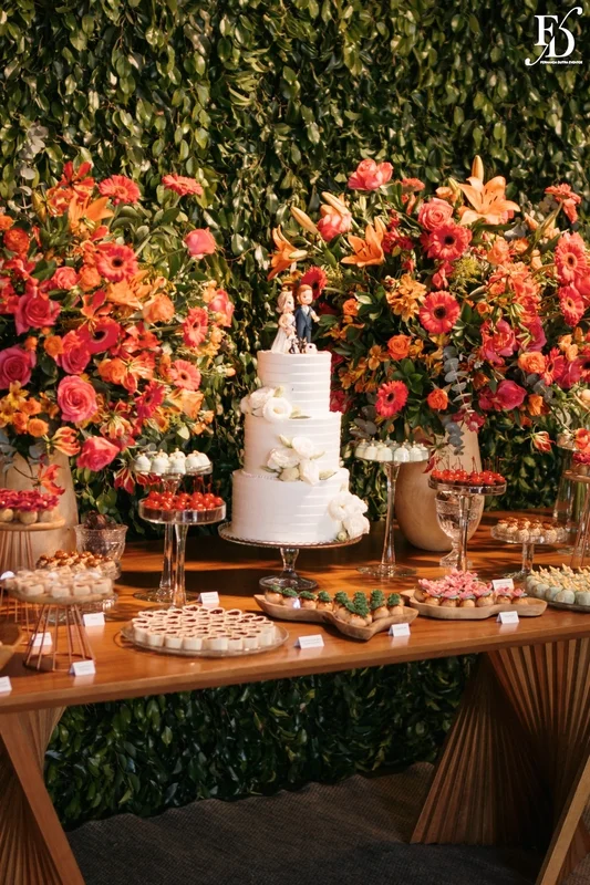 Mesa de doces de casamento com decoração de flores coloridas laranja e rosa e muro inglês