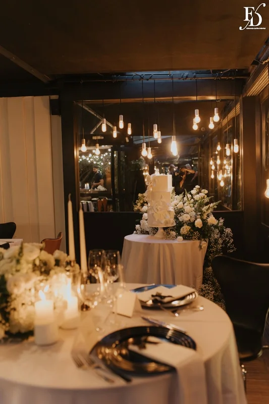 Mesa das noivas com arranjo orgânico branco, velas e bolo de casamento ao fundo no Le Bistrot Gourmet