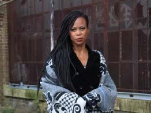 Woman with long braids in a patterned coat stands in front of a brick wall with red graffiti, looking serious.