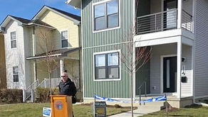 Man speaks at a podium outside green house with "For Sale" sign. Blue sky, neighboring houses visible. Casual, sunny setting.