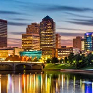 Dusk city skyline with lit office towers and bridge reflected in a calm river under a blue-orange sky.