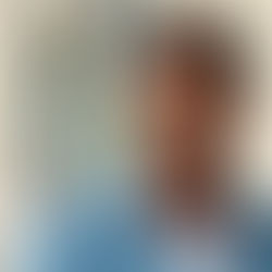 Smiling man in glasses and denim jacket stands by a building. Blurred urban background with warm, inviting colors.