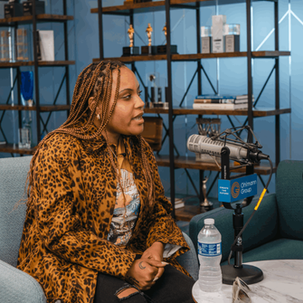 Woman in a leopard-print jacket speaks in a podcast studio with Ohlmann Group mics, blue shelves, trophies, and Create the Future text.