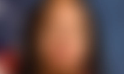 Woman in red blazer poses seriously against a blue background with a partial view of a U.S. flag.