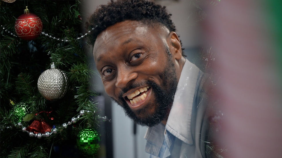 Man smiling near decorated Christmas tree with red, silver, and green ornaments. Festive atmosphere with blurred colorful lights.