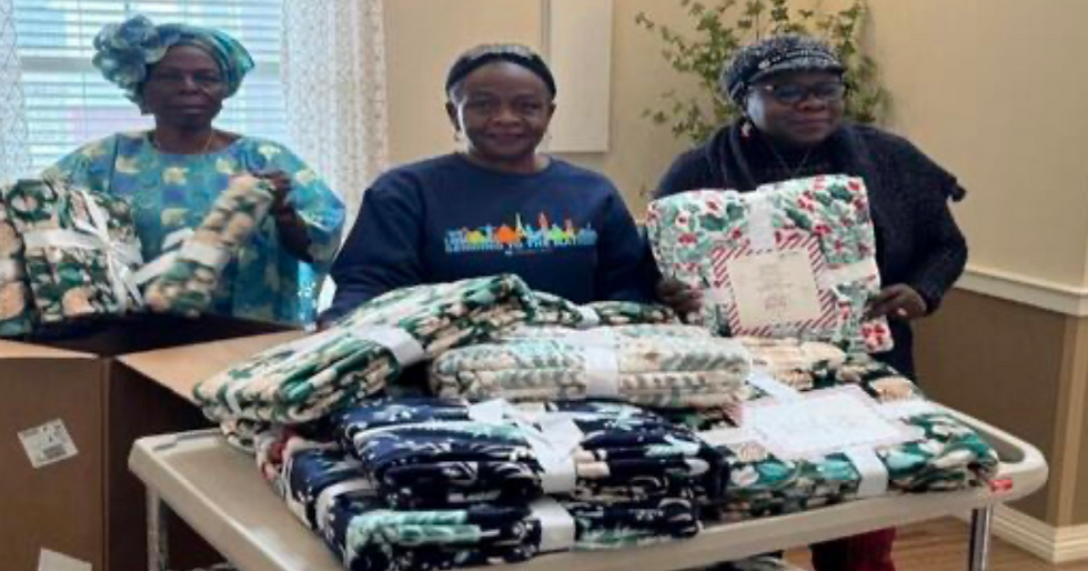 Three women stand behind a table piled with colorful, patterned fabric bundles. One box is open. A joyful mood in a bright room.