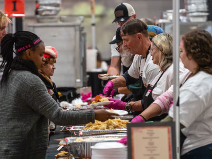 Volunteers wearing white shirts serve food to people at a community event. They stand behind a table with trays in a bustling indoor setting.