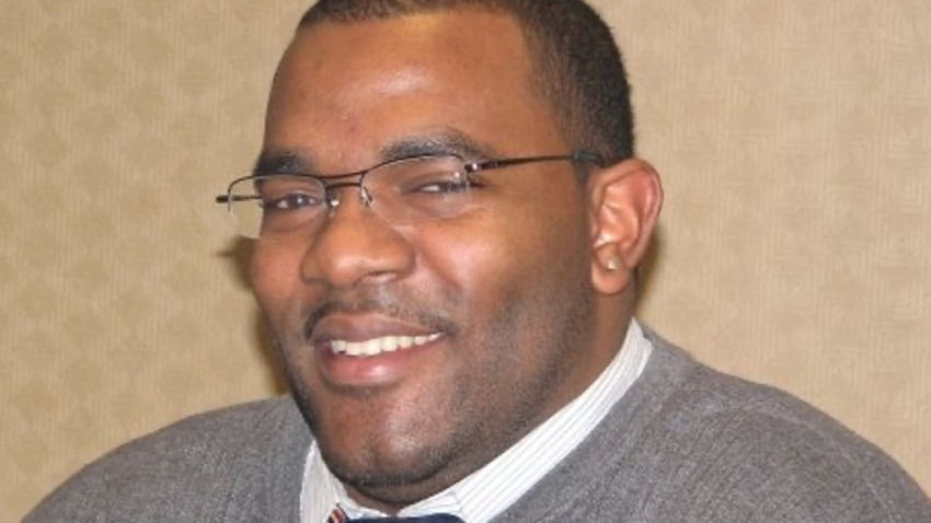 Smiling man in glasses, wearing a sweater and tie, sits in front of a laptop. Beige patterned wall in the background.