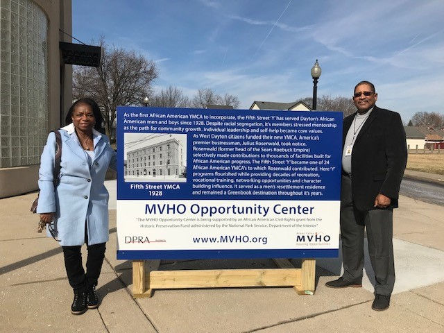Two people stand beside a blue MVHO Opportunity Center sign about Fifth Street YMCA 1928 on a sunny sidewalk.