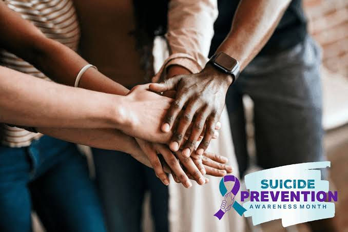 Diverse group stacks hands in support, with Suicide Prevention Awareness Month ribbon logo in the lower right.