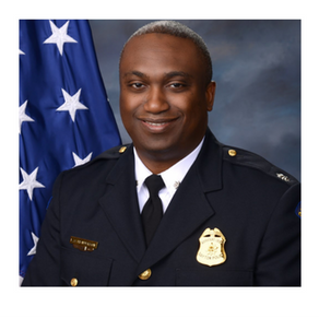 Officer in navy uniform smiling in front of a blue flag with white stars. The badge reads "Assistant Chief Dayton Police."