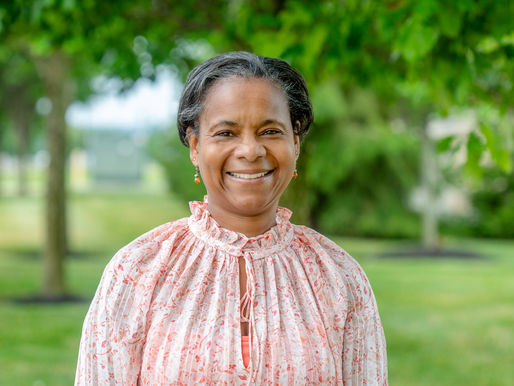 Smiling woman in a pink floral blouse stands outdoors in a green park with blurred trees and grass.