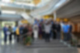 Large group posing on stairs in a bright modern atrium, with yellow accents and glass railings.
