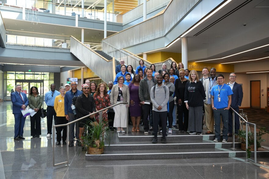 Large group posing on stairs in a bright modern atrium, with yellow accents and glass railings.