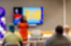 Woman in orange speaks to seated audience at a conference; screen shows Traci Martin and agenda on housing in Black America.