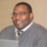 Smiling man in glasses, wearing a sweater and tie, sits in front of a laptop. Beige patterned wall in the background.