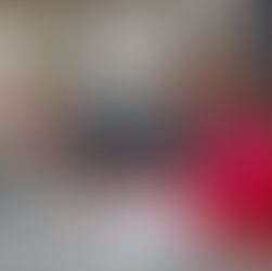 Two women in a library sit at a table, smiling, using a laptop. One points at the screen. Bookshelves are in the background.