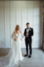 Bride in white off-shoulder dress stands with hands on hips. Groom in black tux adjusts cuff. White paneled wall background.