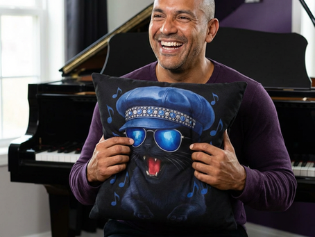 A man is holding a square pillow showing a black cat wearing a blue leather cabbie hat and matching sunglasses. In the background is a piano.