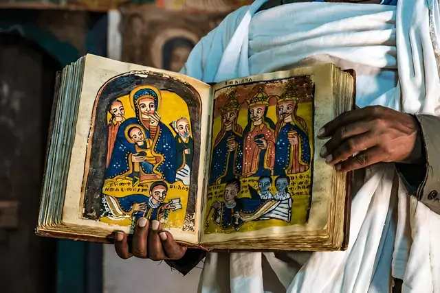 A person in a white robe holds open a colorful biblical manuscript with painted saints and small script.