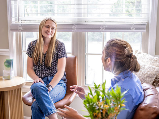 Two women therapists chat in a Life's Journey Counseling's new La Grange Suite 4 office; One smiles in a chair sitting front of a window while they other woman therapist is talking.