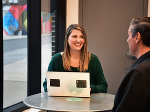 Emily, owner of Small Business Startup Solutions and Wix website designers is smiling at a small business owner in a cafe in Allentown. She's using a laptop on a round table with a colorful mural visible through large windows. Warm, casual atmosphere.