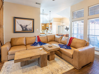 4 Bedroom vacation home in Frisco Texas featuring a modern themed living room that sits off of the kitchen. The Texas themed living room has natural tones with a tan leather sectional, natural wood tables, and a picture of a longhorn above the couch.
