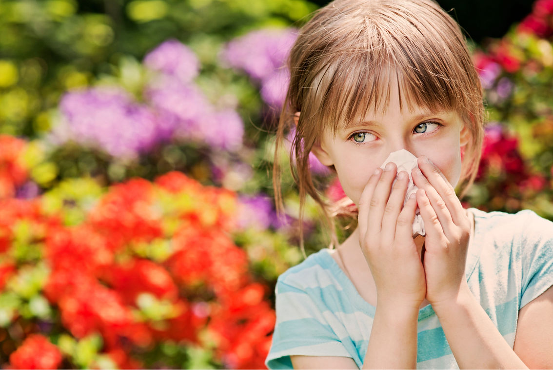 Young girl holding a tissue to her nose, appearing to have allergies, with bright flowers in the background.