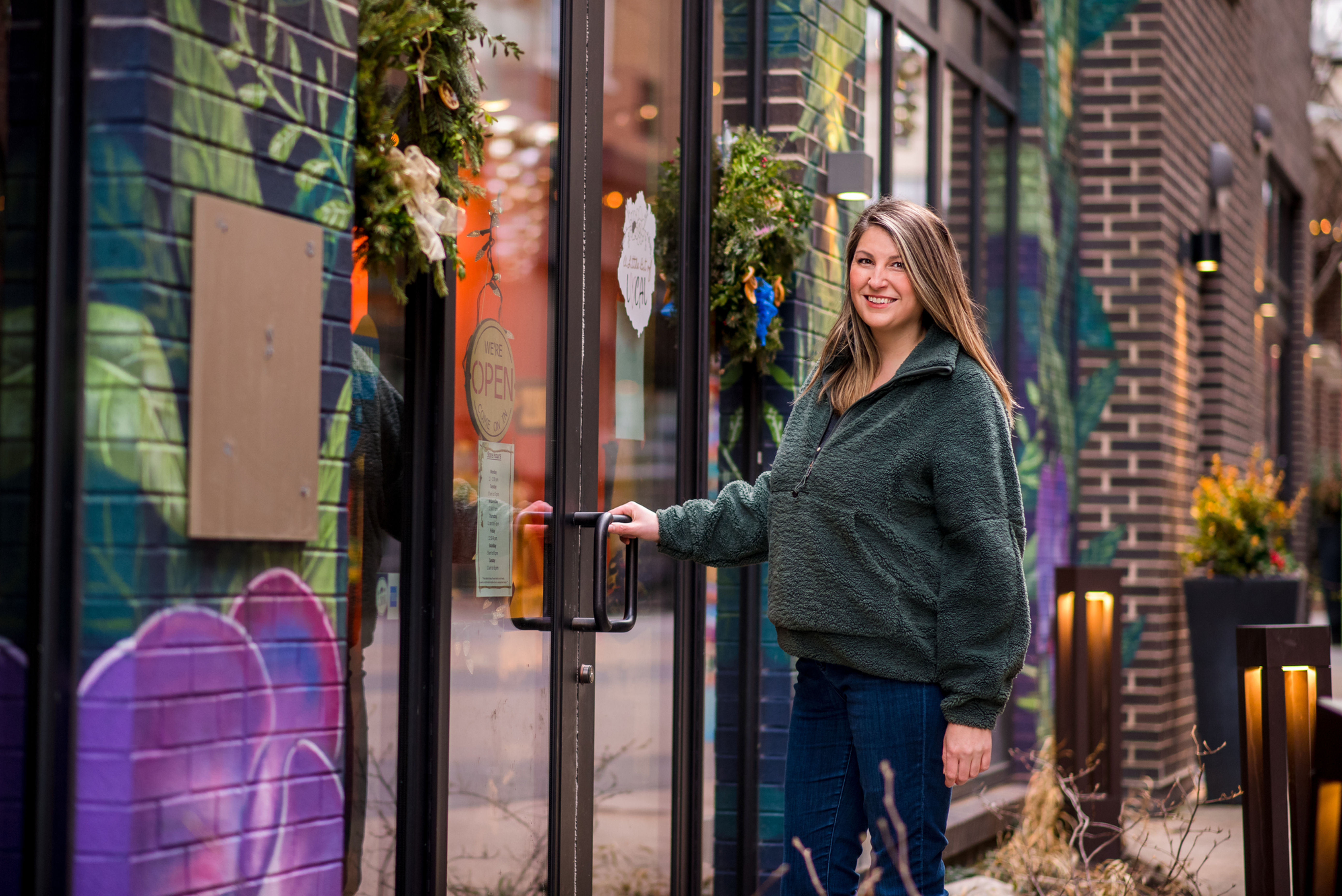 Wix website designer opening the door of a small business in Allentown, PA. She is wearing a green sherpa hoodie, jeans and smiling