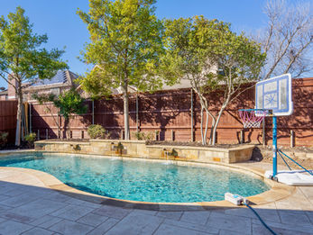 Backyard swimming pool at a vacation rental property in Frisco, Texas featuring a curved in-ground pool, stone water feature, basketball hoop, mature trees, and a private fenced yard under a clear blue sky.