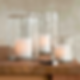 Three lit candles in textured glass holders sit on a wooden surface. A matchbox is nearby. Warm, serene ambiance with soft lighting.