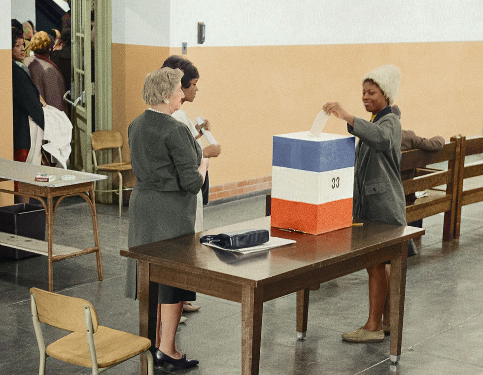 A colorized photo from 1964: A black woman casts her ballot into a red, white and blue box, as several other black women wait in line to cast their own ballots.