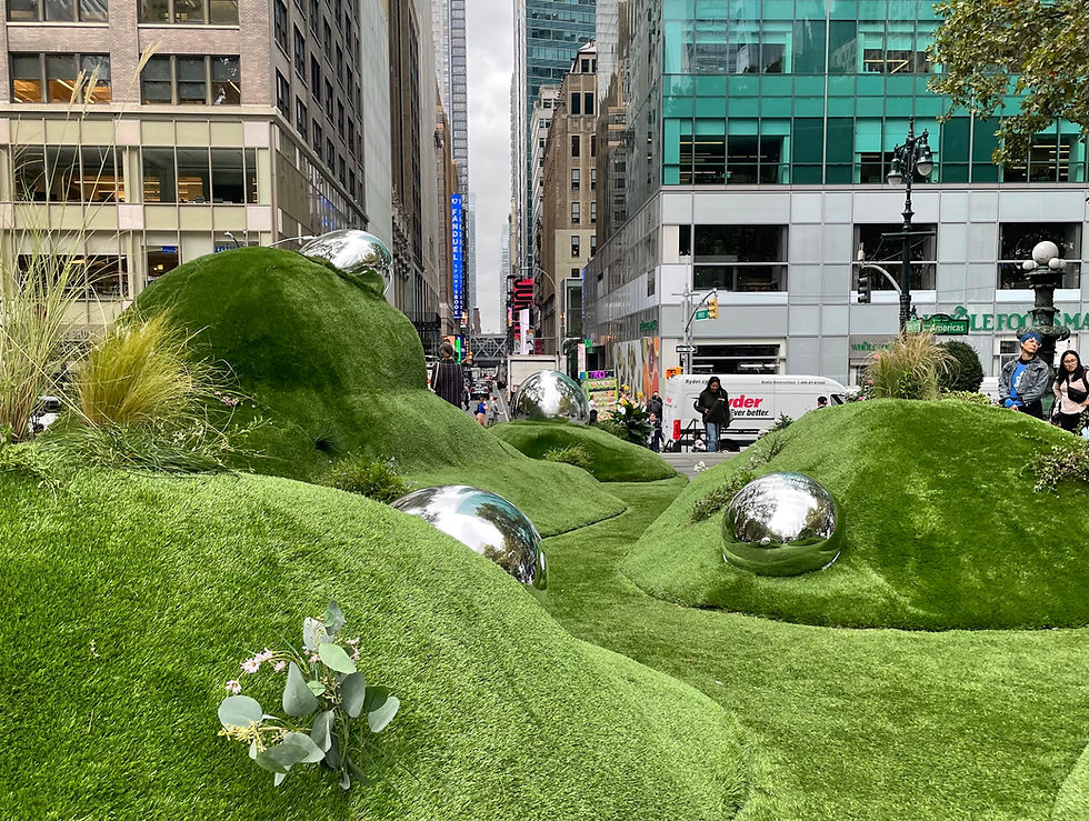Hills covered in astroturf with giant silver balls nestled in the hills. The installation is situated at the end of a canyon of large buildings in Midtown Manhattan.