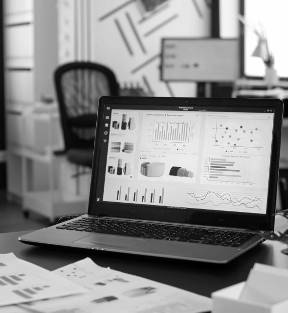 Black and white photo of a laptop on a desk displaying various business analytics dashboards, including bar charts, pie chart