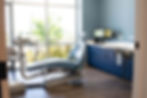 A modern dental examination room featuring a blue dental chair positioned near large floor-to-ceiling windows.