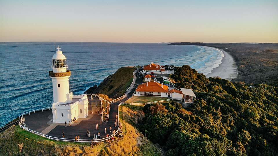 Byron Bay Lighthouse on a cliff at sunset with ocean views, surrounded by people on a walkway; red-roofed buildings and lush greenery nearby.