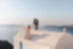 Groom helping the bride up on top of a white building, with the sea and mountains in the background.