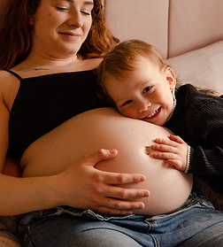 A pregnant lady holding her stomach with her toddler laying his head on her.