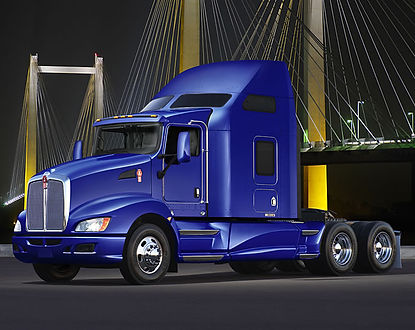 blue semi-truck parked in front of a cable-stayed bridge at night. The truck appears to be a Kenworth model with a sleeper ca