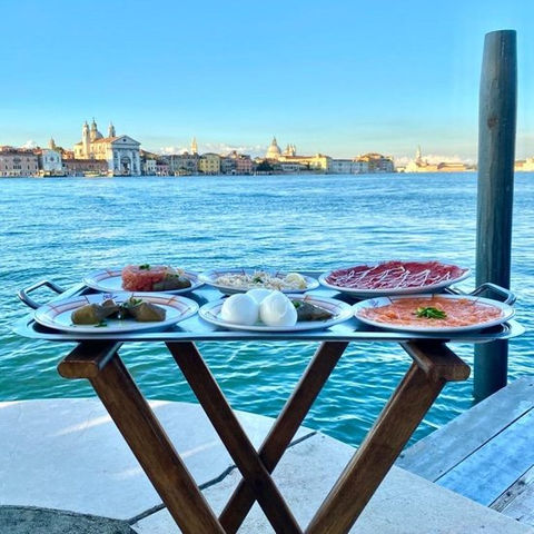 A tray of Cipriani’s iconic Venetian dishes—carpaccio, mozzarella, and smoked salmon—set against a serene waterfront backdrop.