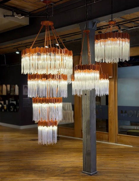 Modern showroom with four tiered hanging chandeliers of white tubes and orange accents over a wood floor.