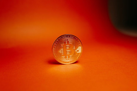 A golden Bitcoin stands on a vivid orange background, showcasing its logo and intricate details. The scene is bright and striking.