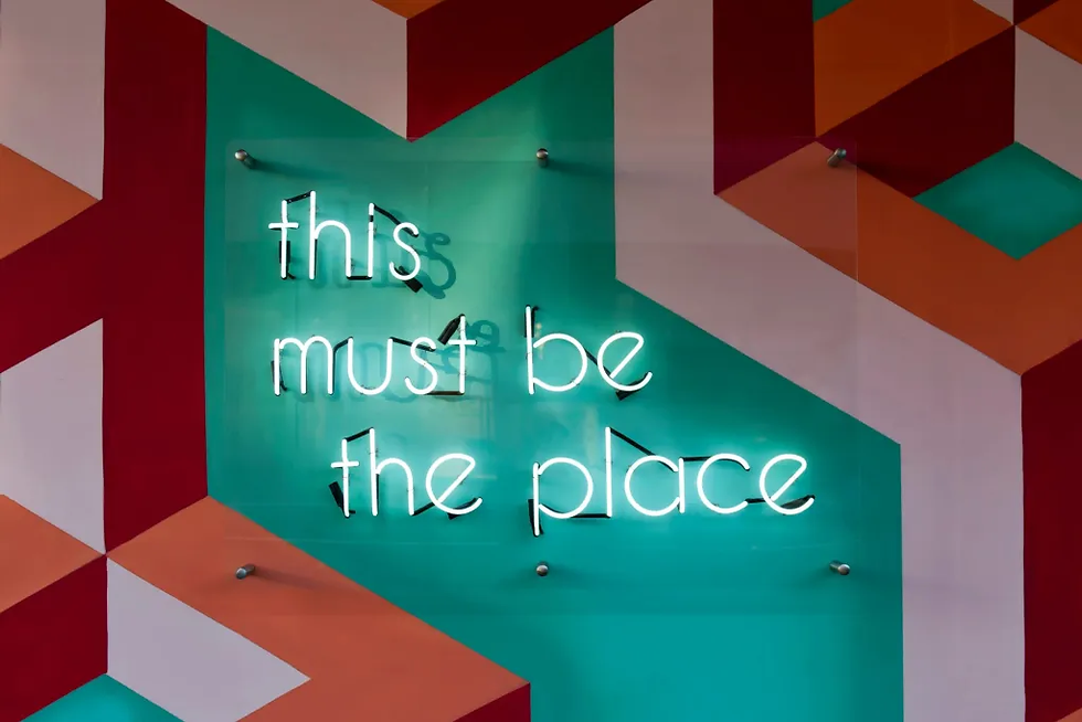 Neon sign reads "this must be the place" on colorful geometric patterned wall with teal, orange, and red shapes. Bright and welcoming mood.