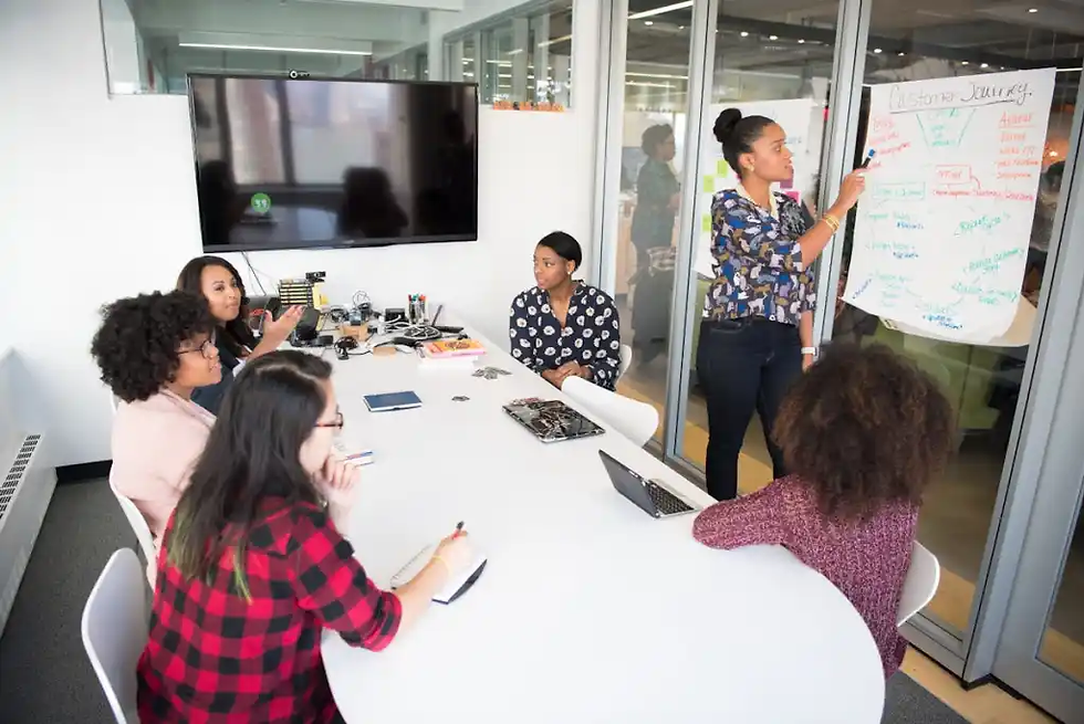 A woman presents at a whiteboard in a modern office. Five seated colleagues listen attentively. The mood is focused and collaborative.