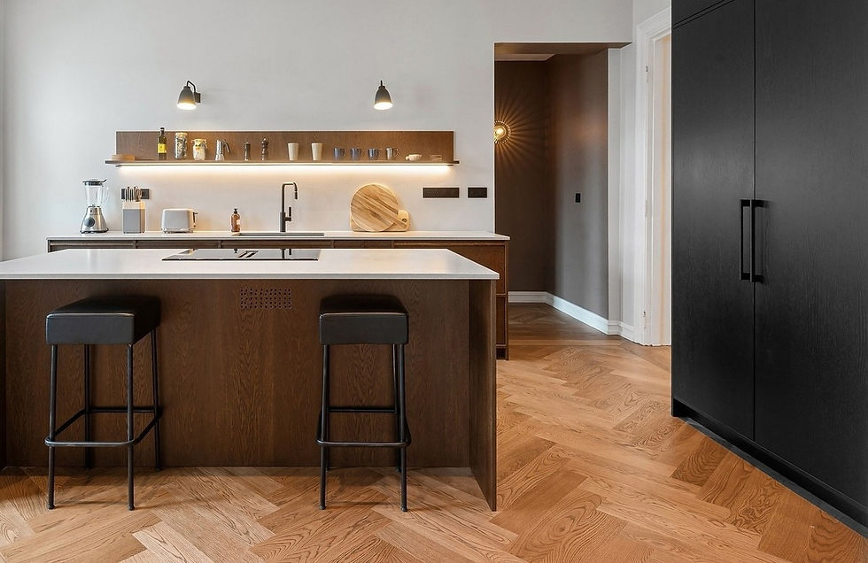 Modern kitchen with dark wood cabinets, white countertops, and black stools. Warm lighting, shelves with cups. Pale herringbone floor adds elegance.