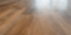 Close-up of light brown wooden floor with a smooth finish and distinct grain patterns. Bright, natural light reflects on the surface.