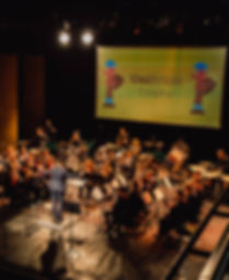 Photo of brass band performing in Theatr Bryn Terfel at a UniBrass contest hosted at Bangor University. Background of 2019 UniBrass Contest logo