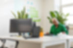 Woman in a green shirt works at a desk with computers. A cactus and plants are in the background. Sticky notes on the wall. Office setting.