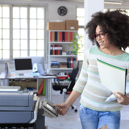 Woman at printer in a bright office, holding papers and smiling. Background has shelves, a desk with computers, and large windows. Relaxed mood.